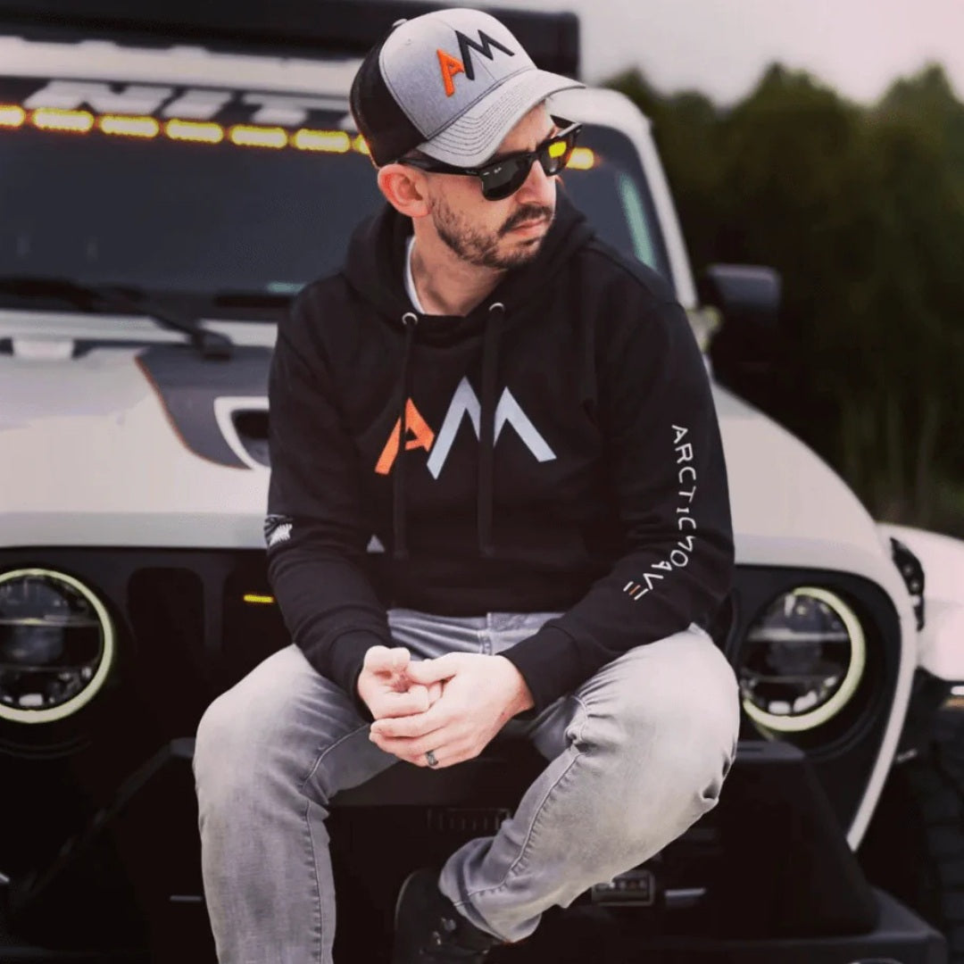 A man wearing an Arctic Move branded black hoodie and matching cap sitting on the front bumper of a white Jeep.