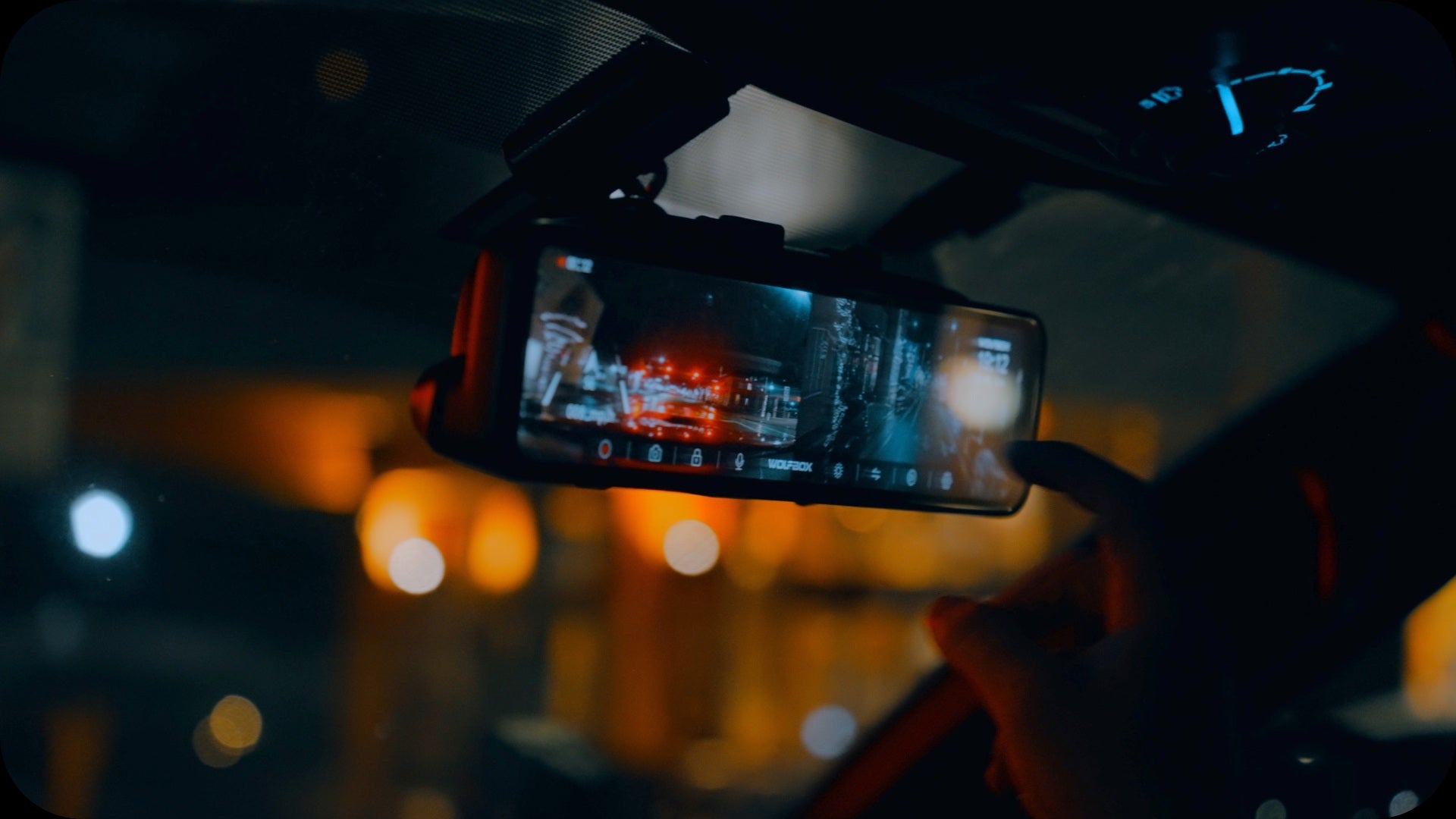 A person operating a Wolfbox rearview mirror dash cam with enhanced night vision display inside a car at night.